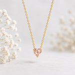 A delicate gold chain necklace with a heart-shaped peach-pink gemstone pendant, displayed against a soft, light gray background with blurred white baby's breath flowers on the sides.
