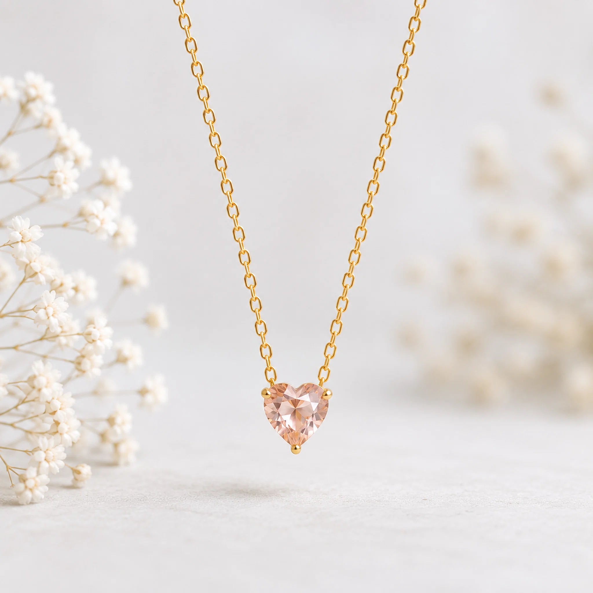 A delicate gold chain necklace with a heart-shaped peach-pink gemstone pendant, displayed against a soft, light gray background with blurred white baby's breath flowers on the sides.