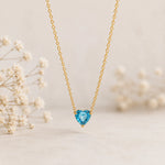 A delicate gold chain necklace with a heart-shaped light blue gemstone pendant, displayed against a soft beige background with blurred white dried flowers on the sides.