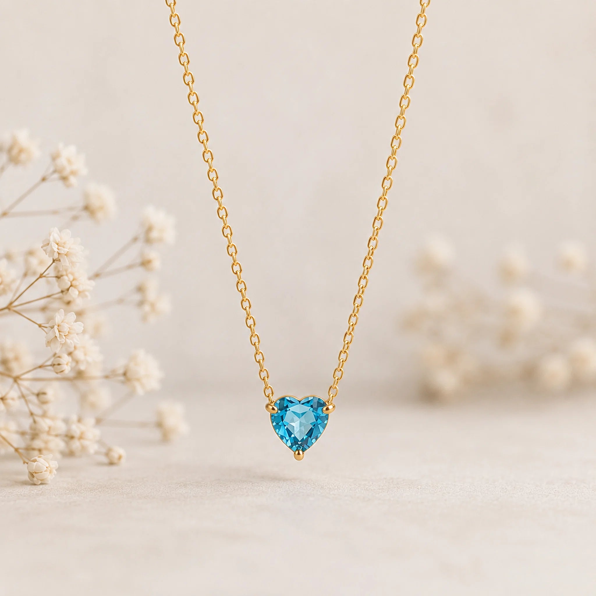 A delicate gold chain necklace with a heart-shaped light blue gemstone pendant, displayed against a soft beige background with blurred white dried flowers on the sides.