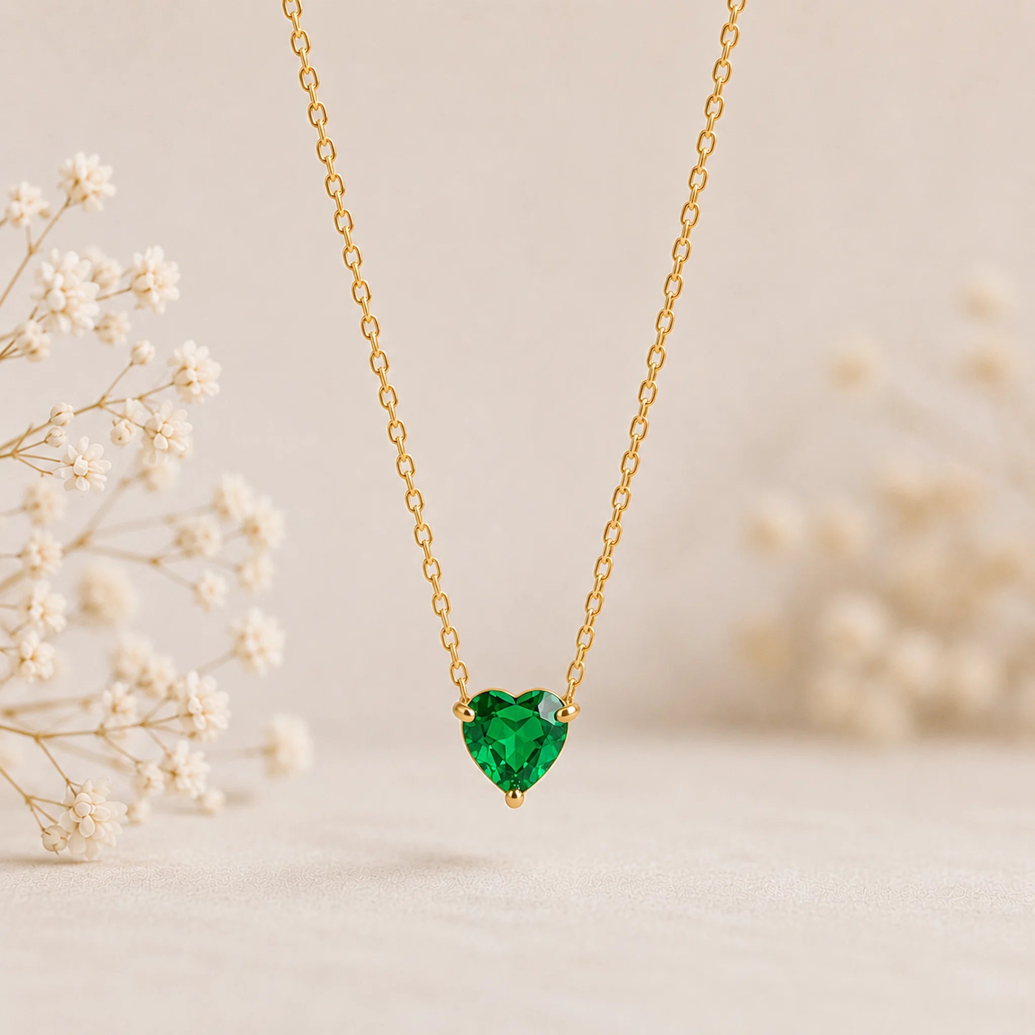 A delicate gold chain necklace with a heart-shaped green gemstone pendant, displayed against a soft beige background with blurred white baby's breath flowers.