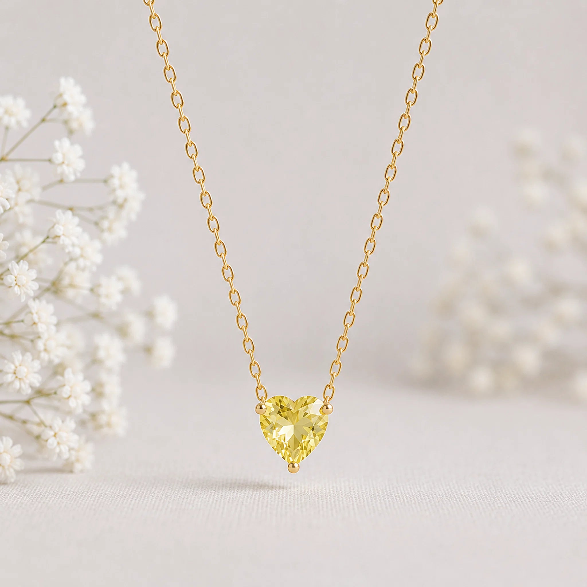 A delicate gold chain necklace with a heart-shaped yellow gemstone pendant, displayed against a soft, light beige background with blurred white baby's breath flowers on the sides.
