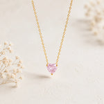 A delicate gold chain necklace with a small, heart-shaped pink gemstone pendant, displayed on a light textured surface with soft, blurred dried flowers in the background.