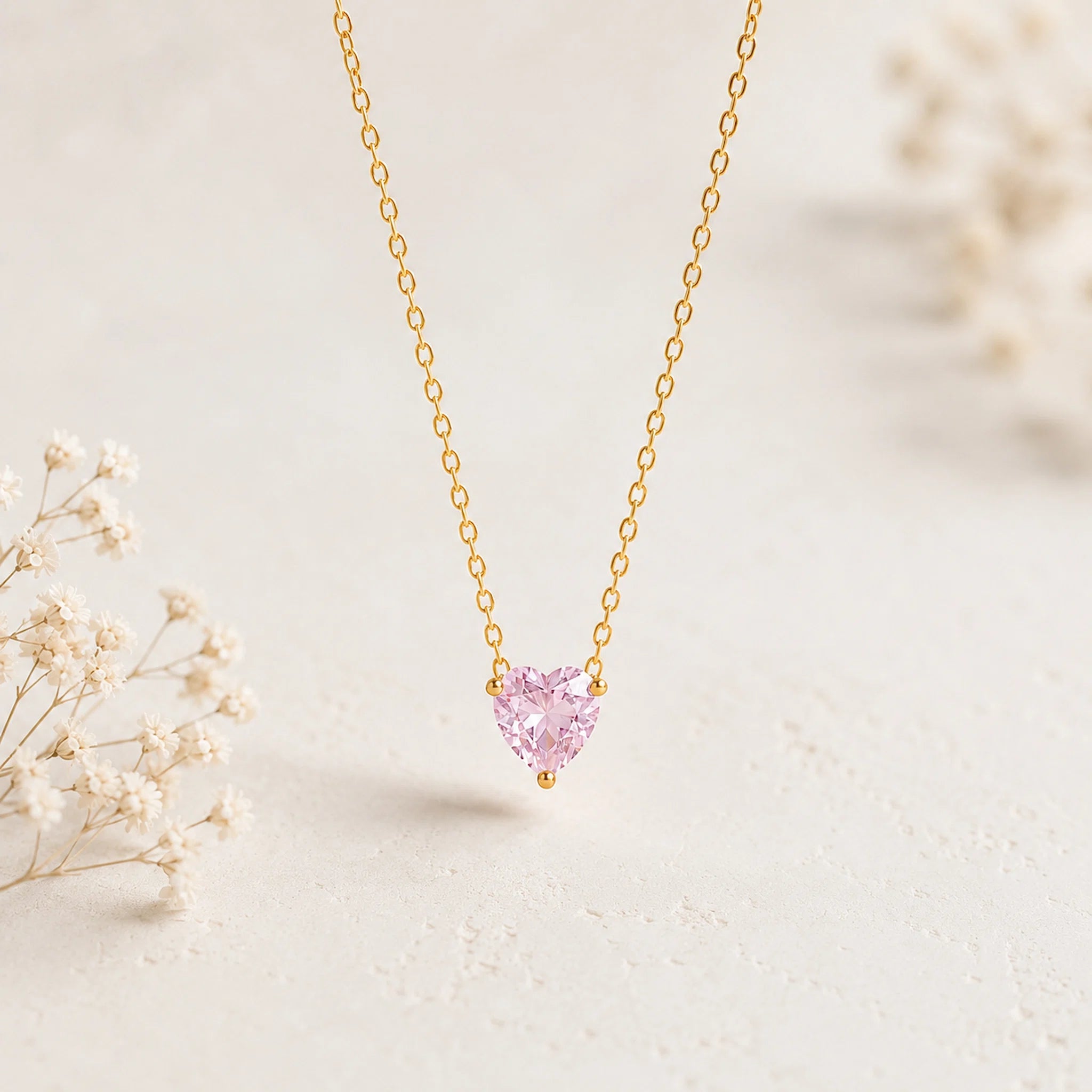A delicate gold chain necklace with a small, heart-shaped pink gemstone pendant, displayed on a light textured surface with soft, blurred dried flowers in the background.