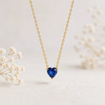 A delicate gold chain necklace with a heart-shaped deep blue gemstone pendant, displayed against a soft, light background with blurred white baby's breath flowers.