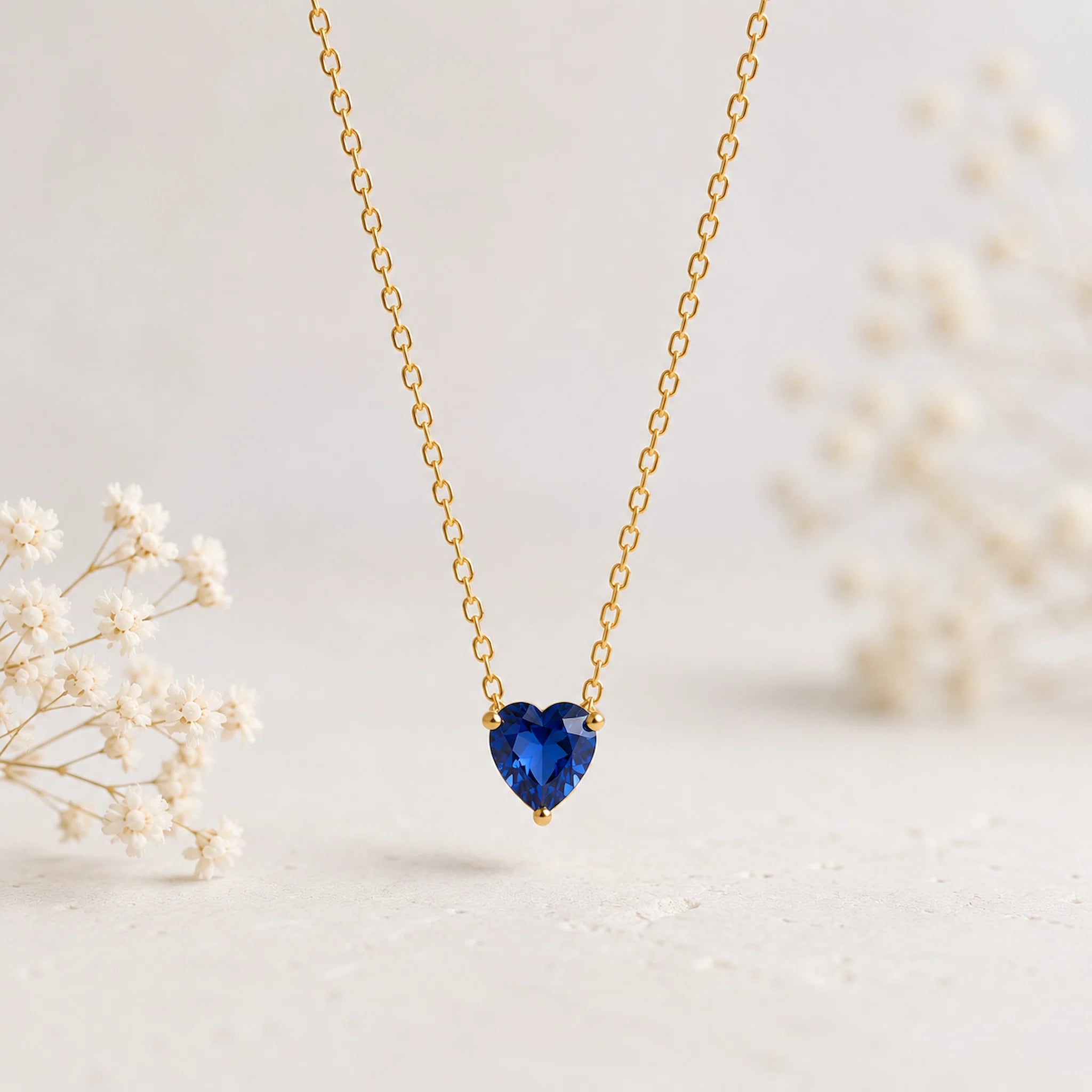 A delicate gold chain necklace with a heart-shaped deep blue gemstone pendant, displayed against a soft, light background with blurred white baby's breath flowers.