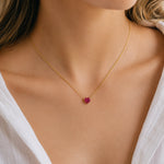 A delicate gold chain necklace with a small, heart-shaped pink gemstone pendant, worn by a model in a white top.