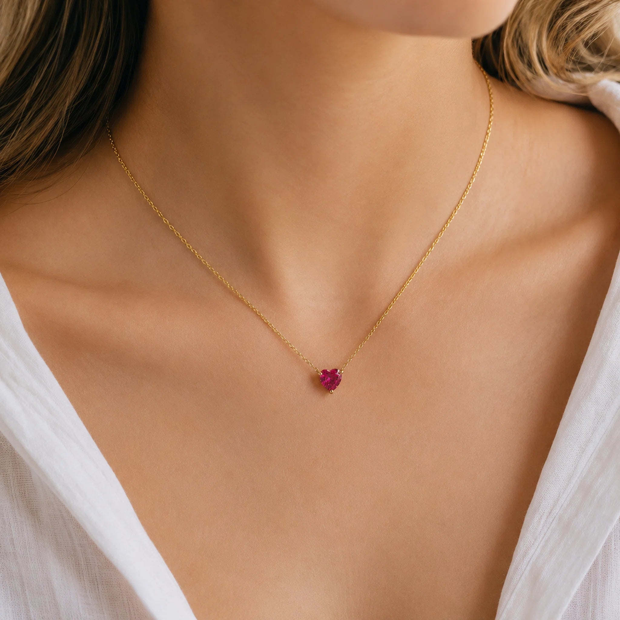 A delicate gold chain necklace with a small, heart-shaped pink gemstone pendant, worn by a model in a white top.