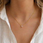 A delicate gold chain necklace with a small heart-shaped clear gemstone pendant, worn by a model in a white blouse.