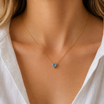 A close-up of a model wearing a delicate gold chain necklace with a small, heart-shaped light blue gemstone pendant, styled with a white collared shirt.