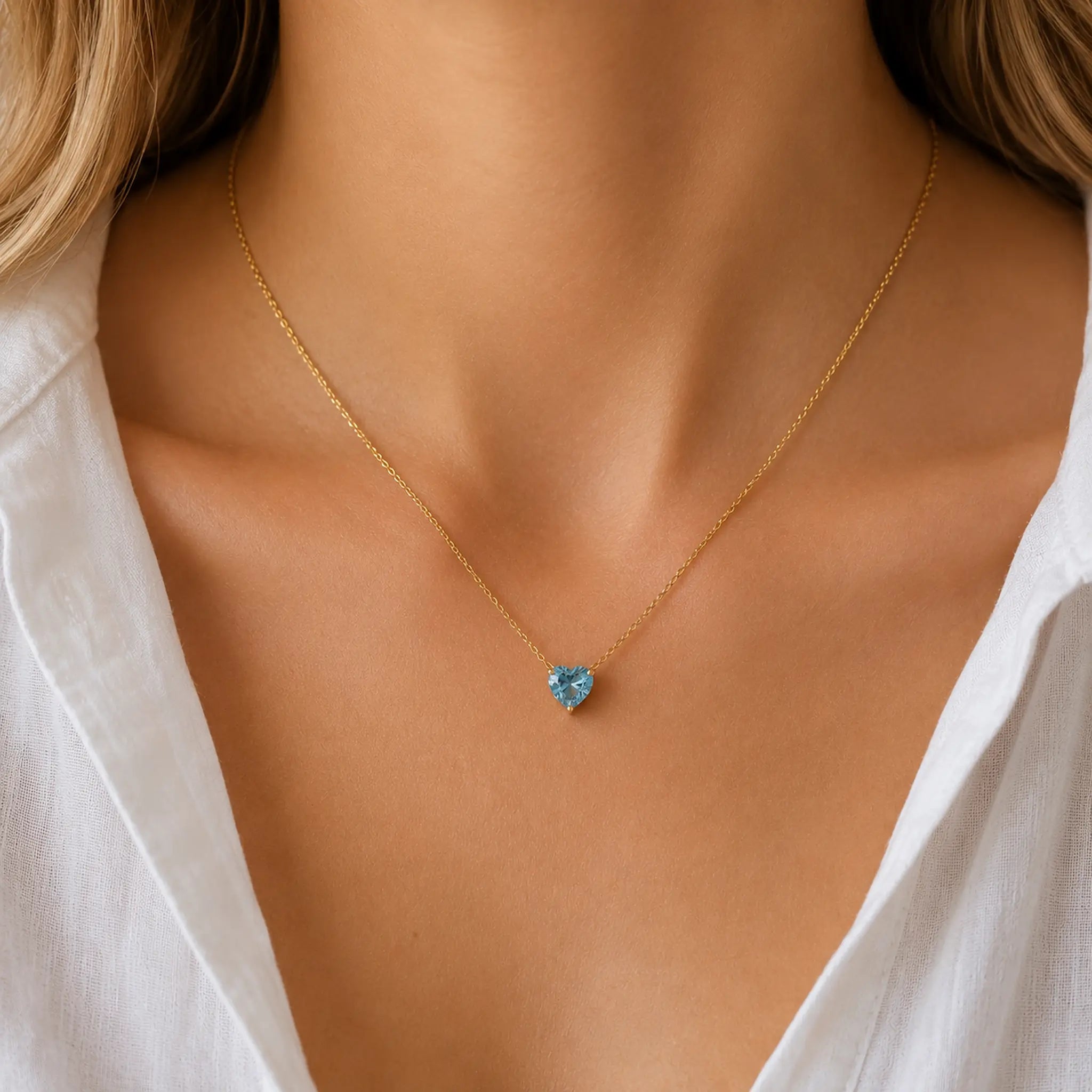 A close-up of a model wearing a delicate gold chain necklace with a small, heart-shaped light blue gemstone pendant, styled with a white collared shirt.
