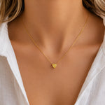 A delicate gold chain necklace with a small textured heart pendant, worn by a model in a white top.