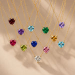 Gold heart birthstone necklaces with 12 heart-cut cubic zirconia colour options hanging on fine gold chains against a warm beige background