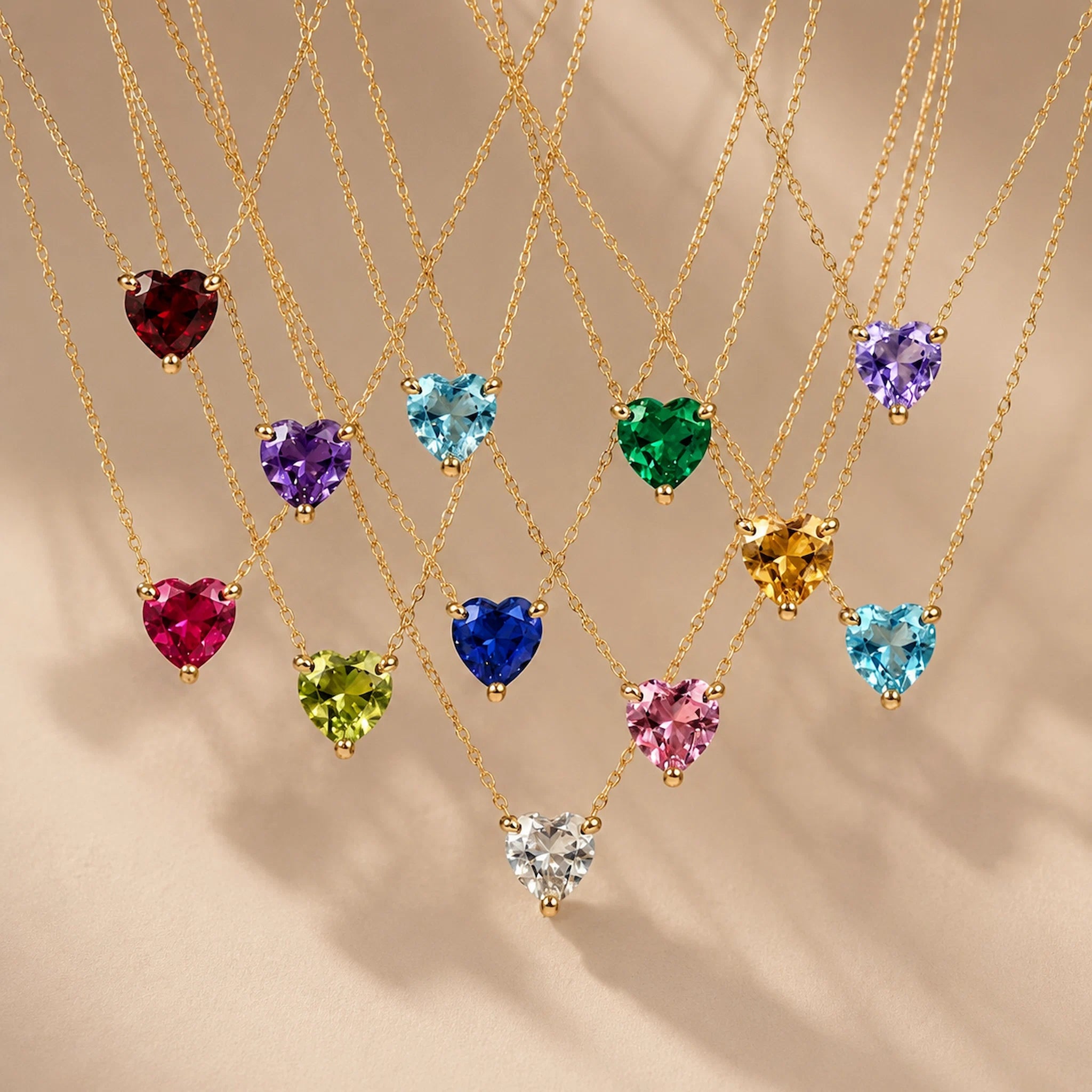 Gold heart birthstone necklaces with 12 heart-cut cubic zirconia colour options hanging on fine gold chains against a warm beige background