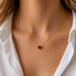 A close-up of a model wearing a delicate gold chain necklace with a small, heart-shaped red gemstone pendant, styled with a white collared shirt.