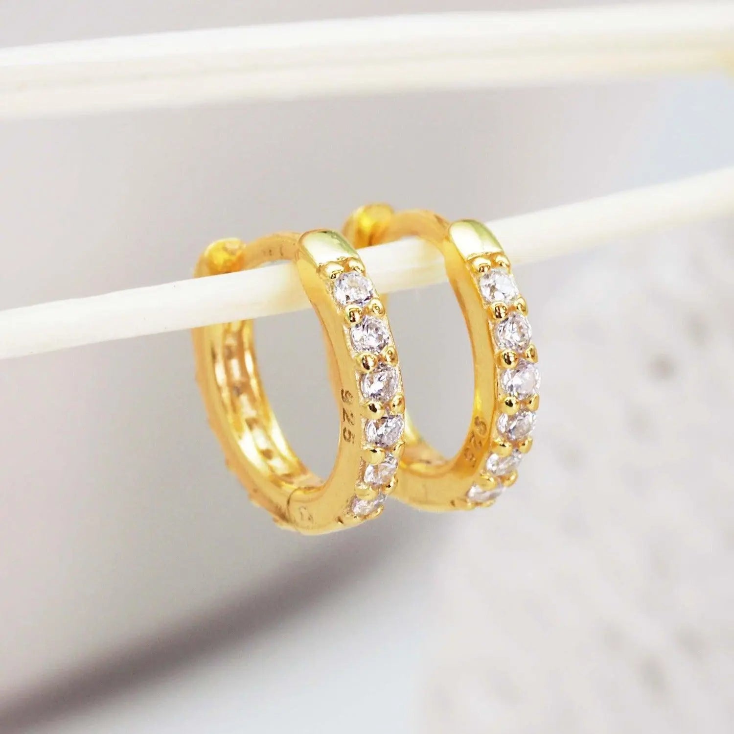Gold Huggie Hoop Earrings with clear cubic zirconias hanging from a white pole