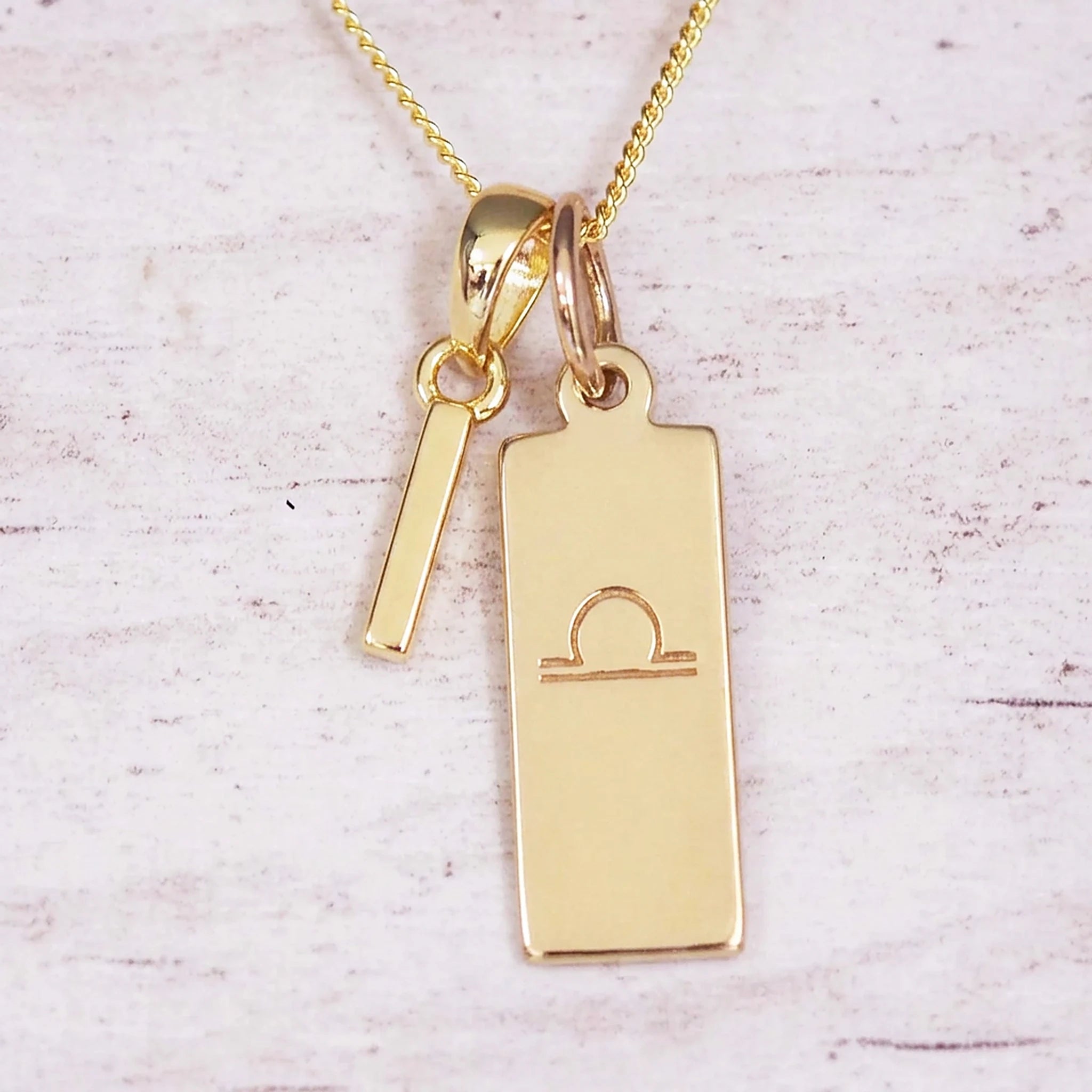 Gold initial and Libra zodiac necklace with a letter I charm and engraved bar pendant on whitewashed wood