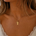 Close-up of a person wearing a gold double pendant necklace with a capital letter pendant and a rectangle pendant with the cancer zodiac glyph.