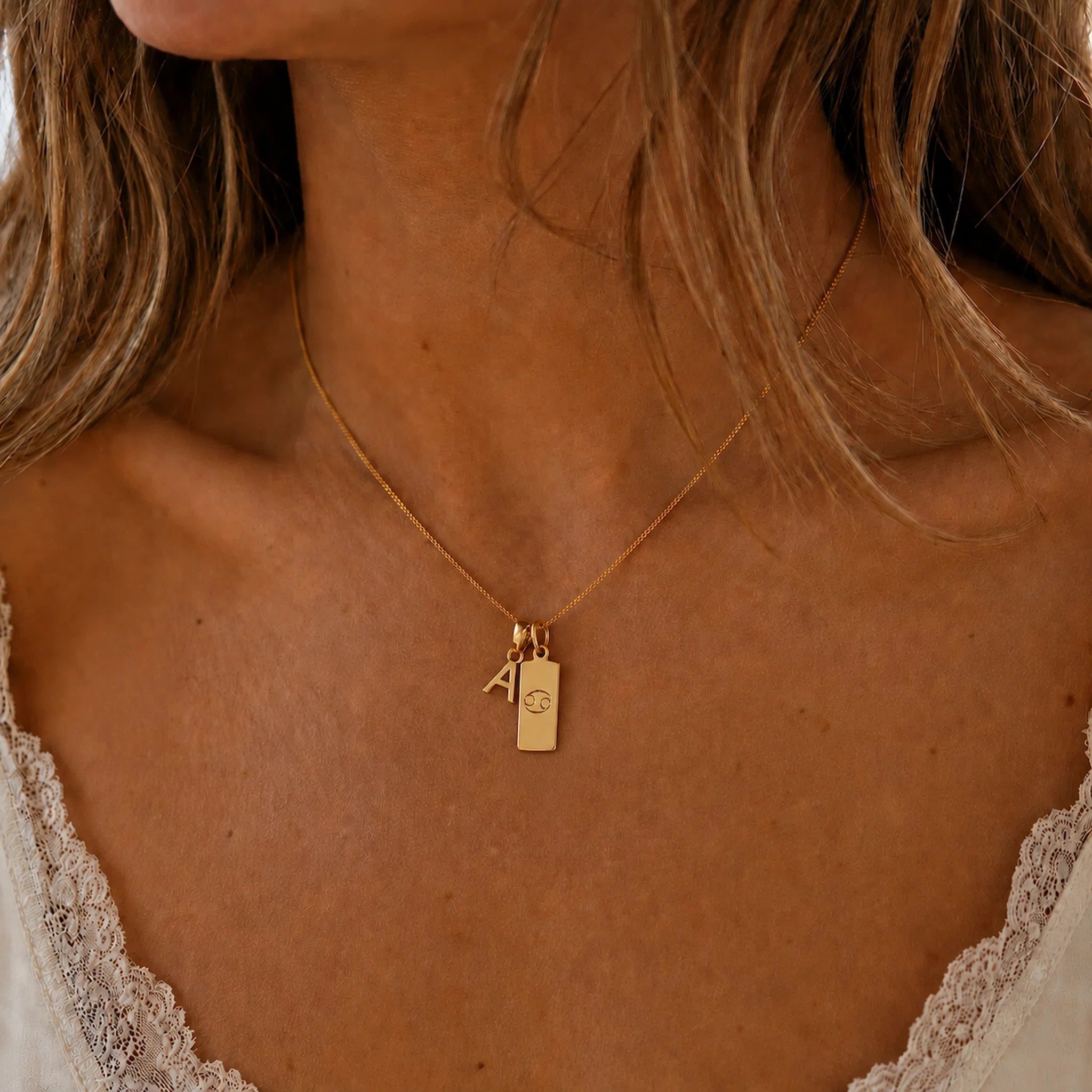 Close-up of a person wearing a gold double pendant necklace with a capital letter pendant and a rectangle pendant with the cancer zodiac glyph.