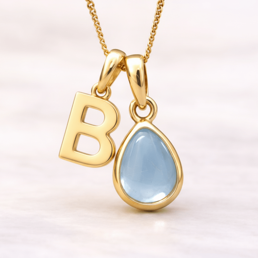 Gold necklace with a 'B' pendant and teardrop-shaped blue topaz gemstone on a light pink background