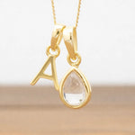Gold necklace with a teardrop-shaped herkimer quartz crystal pendant and letter 'A' charm on a light background