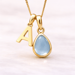 Gold necklace with a teardrop-shaped blue topaz pendant and letter 'A' on a light pink background