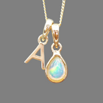 Gold pendant with 'A' and teardrop-shaped gemstone on a gray background