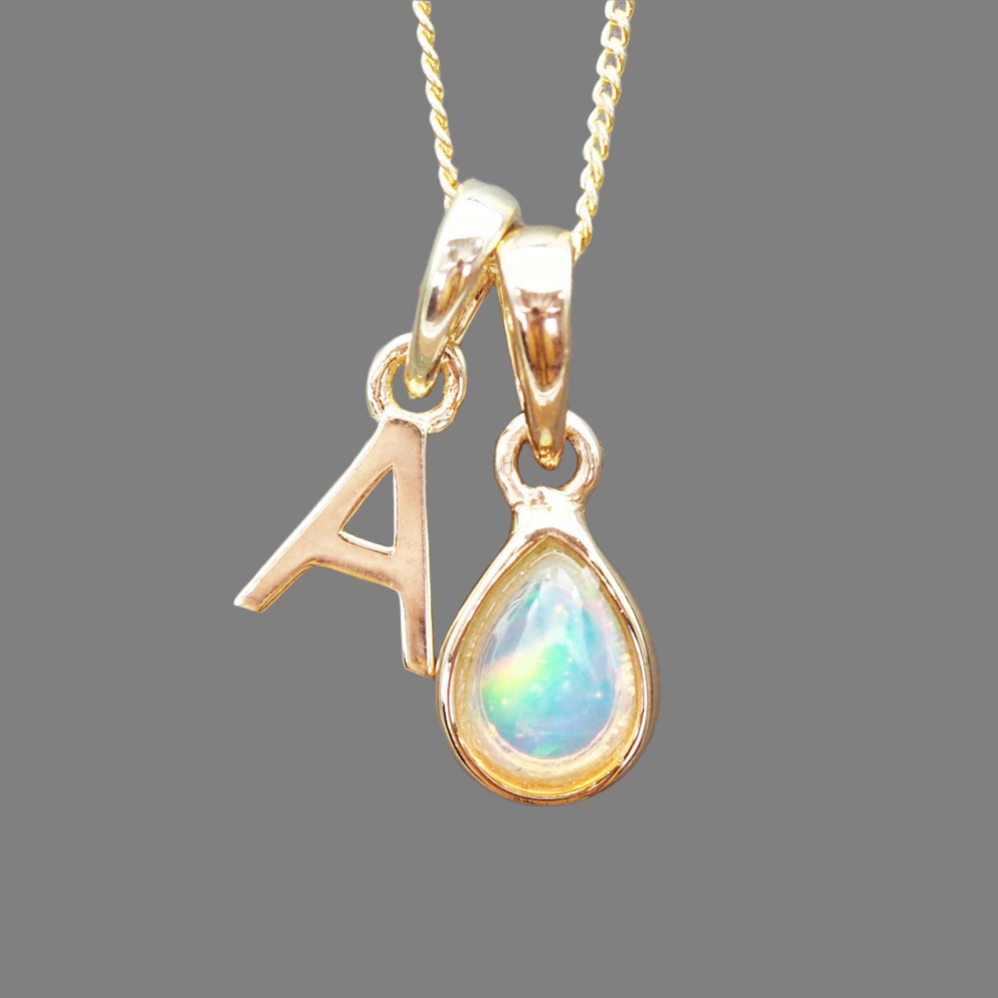 Gold pendant with 'A' and teardrop-shaped gemstone on a gray background