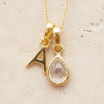 Gold fine chain necklace with 'A' letter initial pendant and a april birthstone pear-shaped clear quartz pendant on a beige background