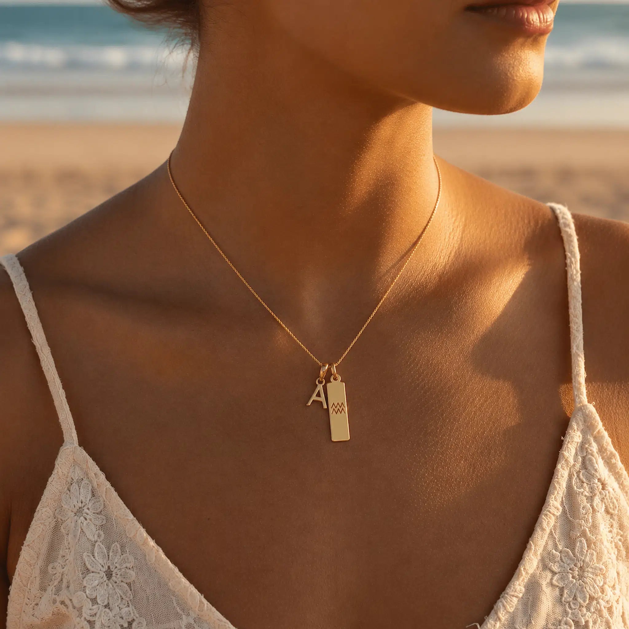 Woman wearing a gold fine chain necklace with a uppercase A letter pendant and a rectangle pendant with the aquarius star sign glyph symbol, on a beach