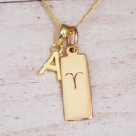 Gold necklace with a pendant featuring the letter 'A' and an Aries zodiac Star sign pendant on a light wooden background.