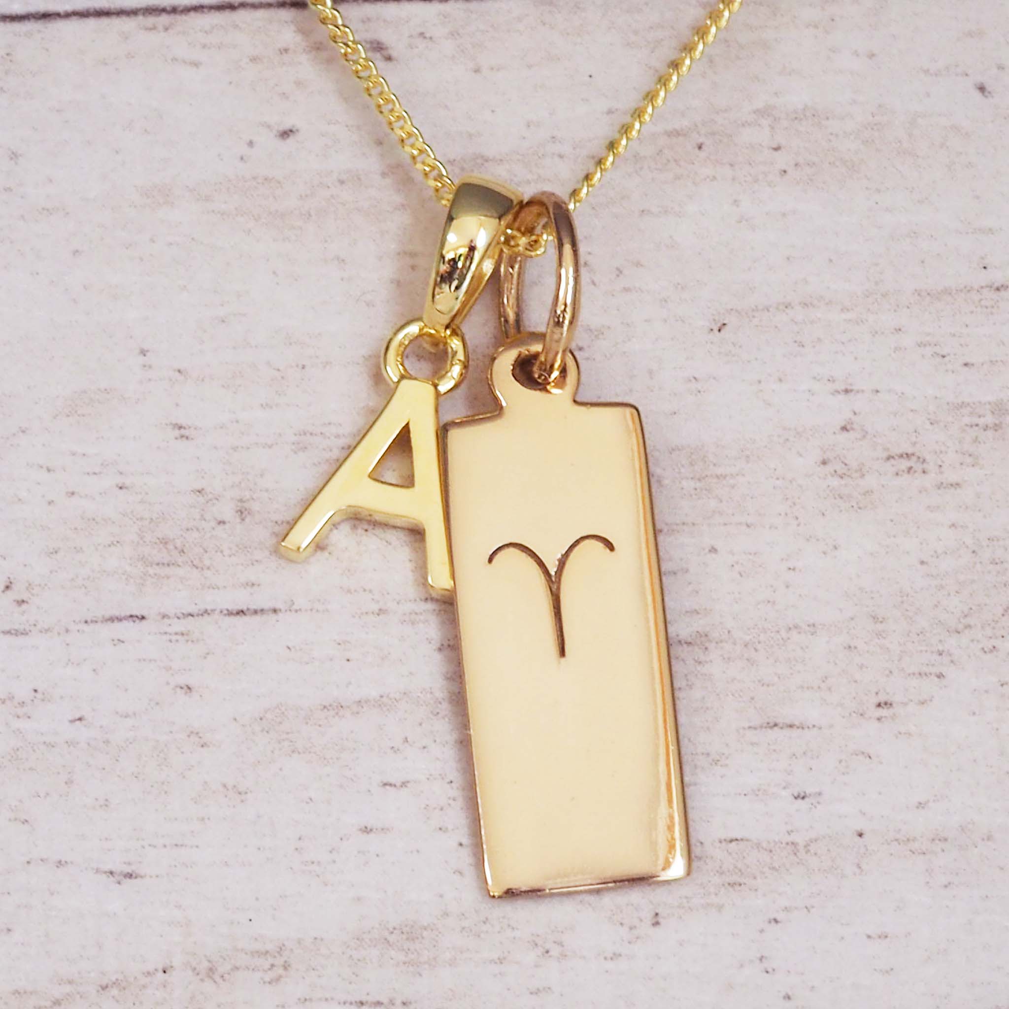 Fine Gold Chain double pendant necklace with a pendant featuring a uppercase letter 'A', and a rectangular pendant with the Aries zodiac Star sign glyph symbol on a light wooden background.