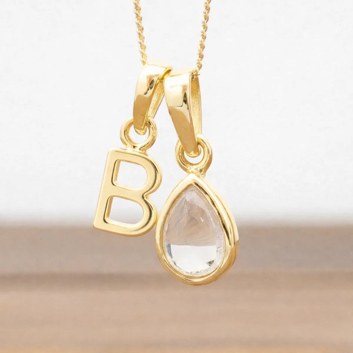 Gold necklace with 'B' pendant and teardrop-shaped herkimer quartz crystal gemstone on a neutral background