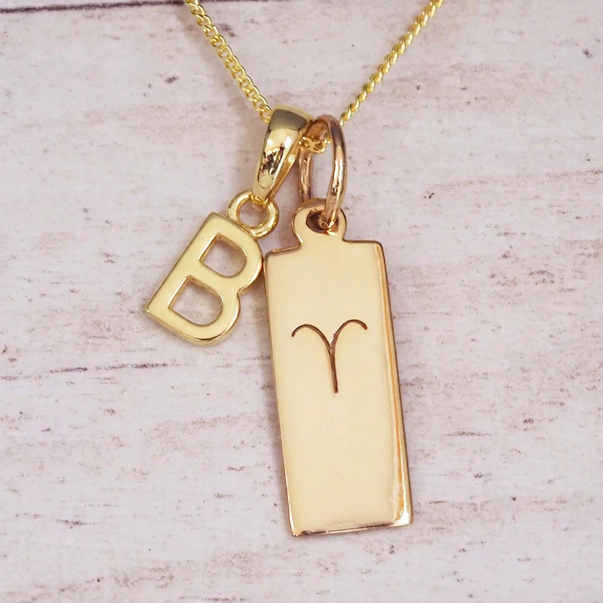 Gold initial B and Aries zodiac bar necklace, close-up of the letter charm and engraved pendant on whitewashed wood