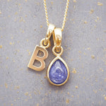 Gold December birthstone necklace with a pear-shaped natural tanzanite pendant and letter 'B' charm with a grey background.