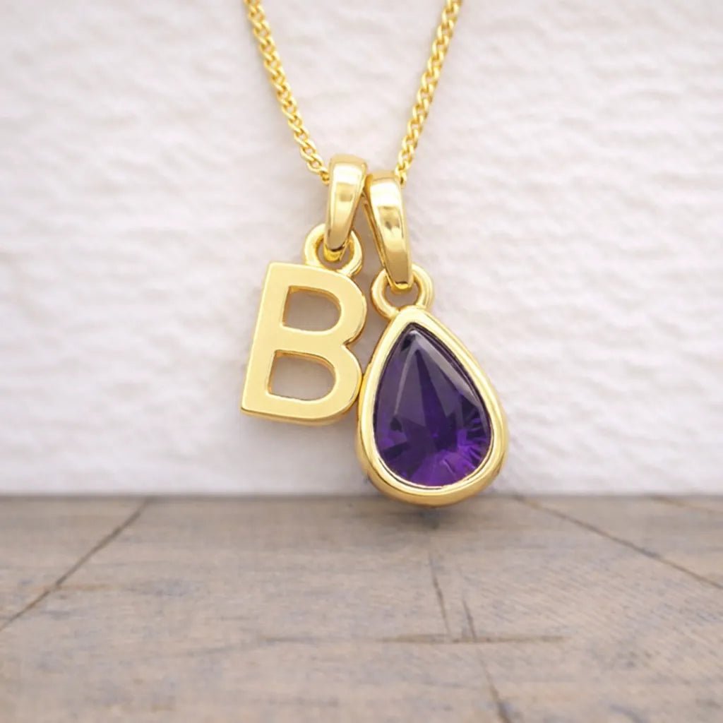 Gold necklace with 'B' letter and teardrop-shaped purple gemstone pendant on a neutral background