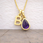 Gold necklace with 'B' letter and teardrop-shaped purple gemstone pendant on a neutral background