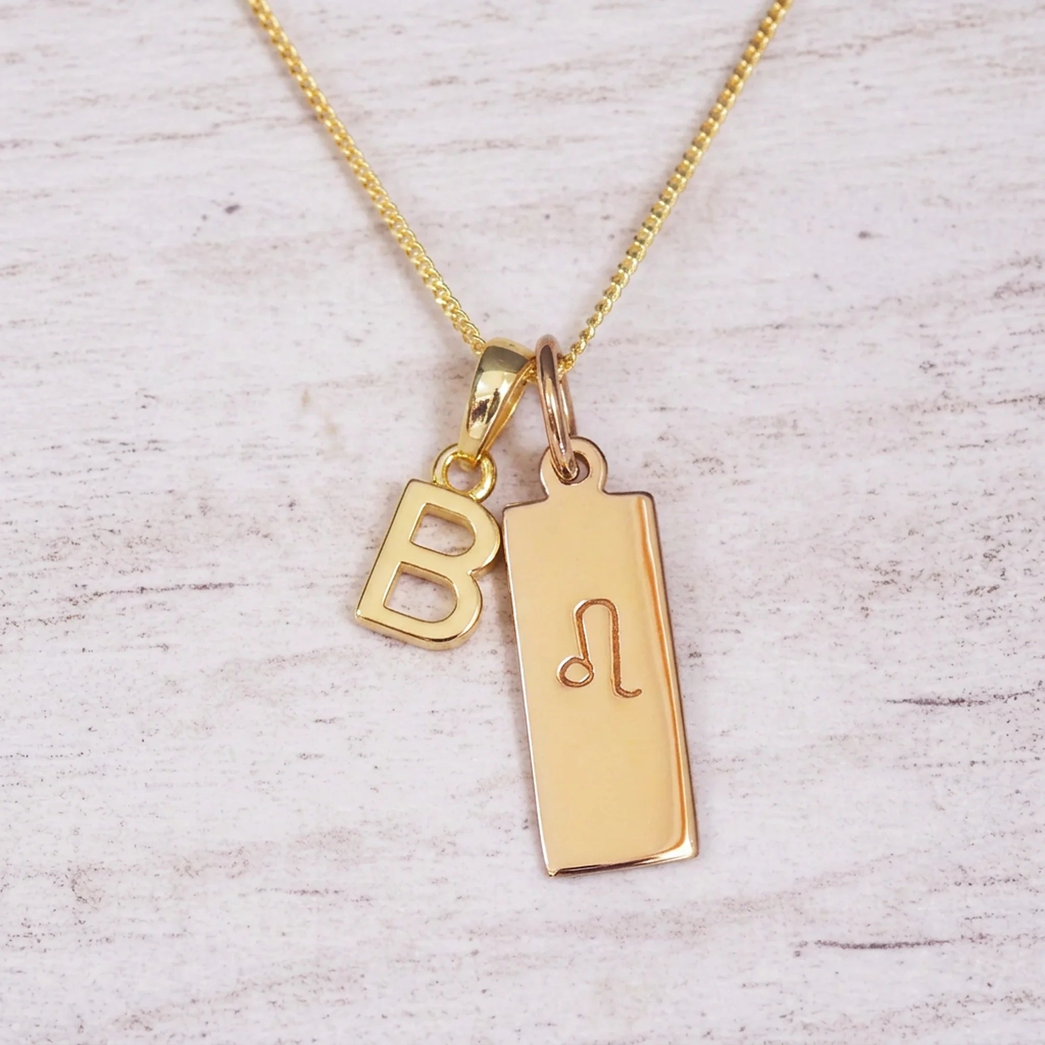 Gold initial and Leo zodiac necklace with a letter B pendant and engraved Leo bar on white wood, close-up