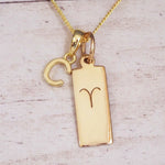 Gold initial C and Aries zodiac bar necklace, close-up of the letter charm and engraved pendant on whitewashed wood