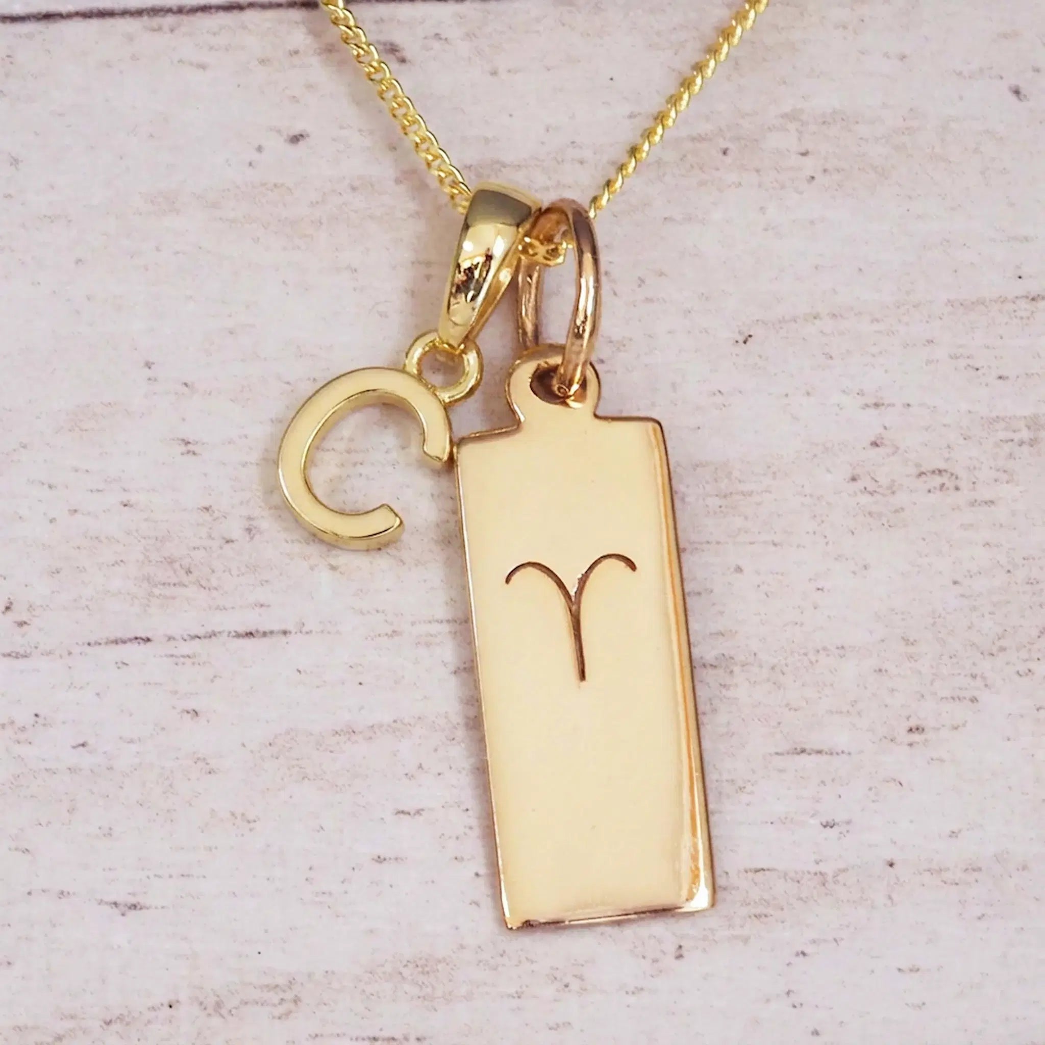 Gold initial C and Aries zodiac bar necklace, close-up of the letter charm and engraved pendant on whitewashed wood