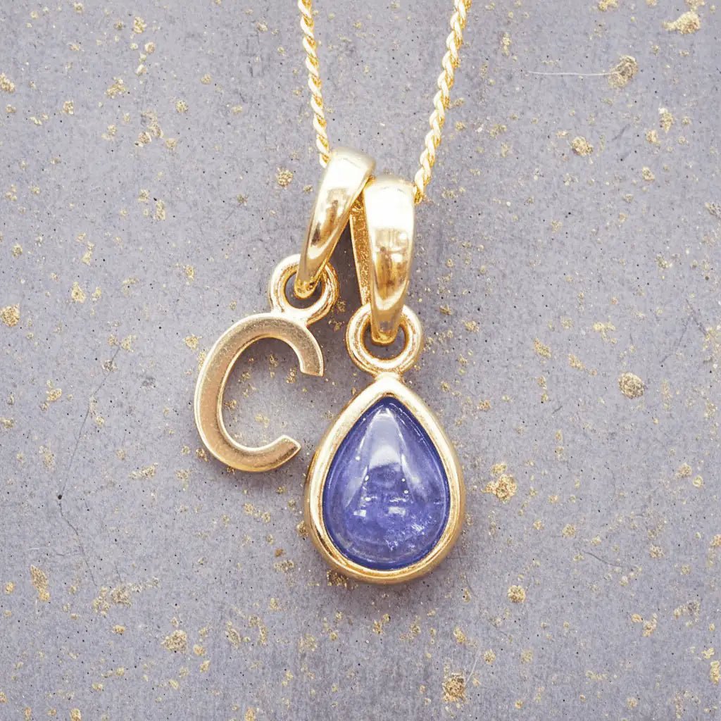 Gold December birthstone necklace with a pear-shaped natural tanzanite pendant and letter 'C' charm with a grey background.