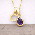 Gold necklace with a 'C' pendant and purple teardrop gemstone on a light background
