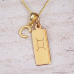 Gold initial C and Gemini zodiac bar necklace, close-up of the letter charm and engraved pendant on whitewashed wood