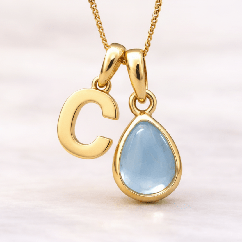 Gold necklace with a 'C' pendant and a teardrop-shaped blue topaz gemstone on a light pink background.