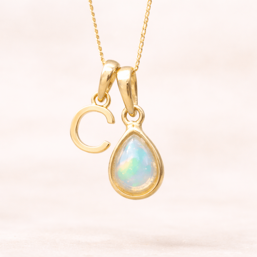 Gold necklace with a C initial pendant and teardrop-shaped opal pendant on a light pink background