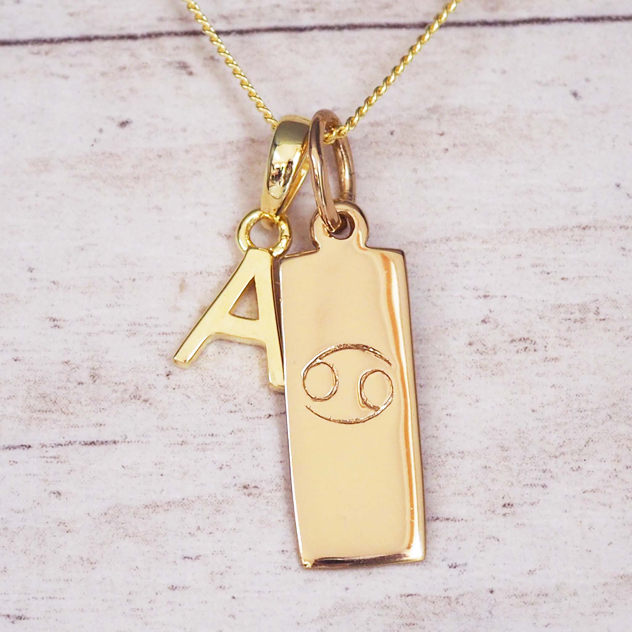 Gold necklace with a pendant featuring the letter 'A' and a cancer zodiac symbol on a light wooden background.