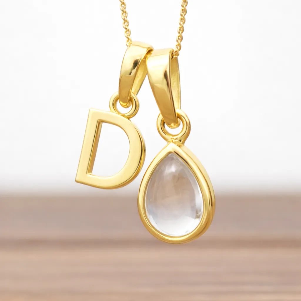Gold pendant with 'D' and teardrop-shaped herkimer quartz crystal gemstone on a gradient background