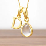 Gold pendant with 'D' and teardrop-shaped herkimer quartz crystal gemstone on a gradient background