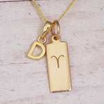Gold initial D and Aries zodiac bar necklace, close-up of the letter charm and engraved pendant on whitewashed wood