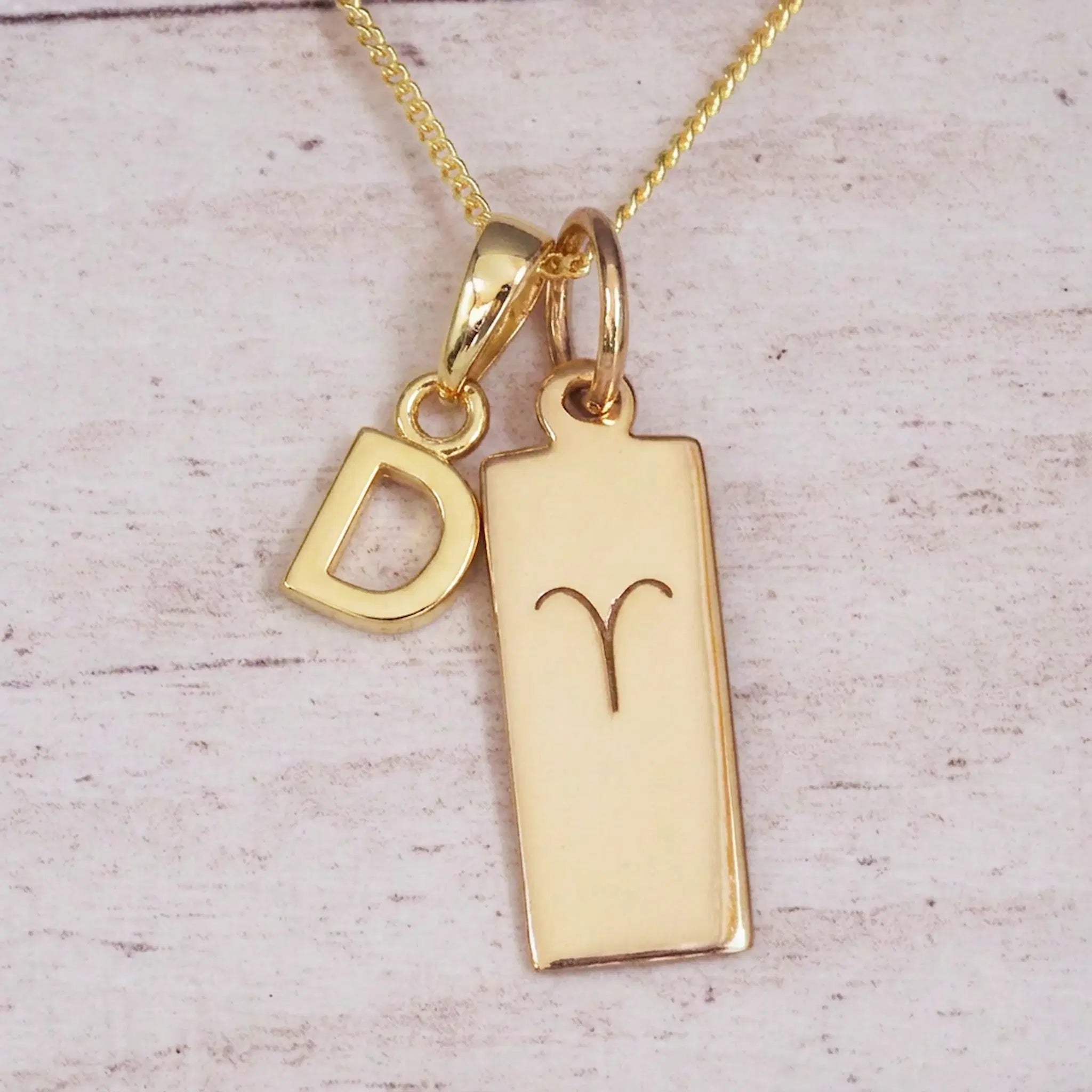Gold initial D and Aries zodiac bar necklace, close-up of the letter charm and engraved pendant on whitewashed wood